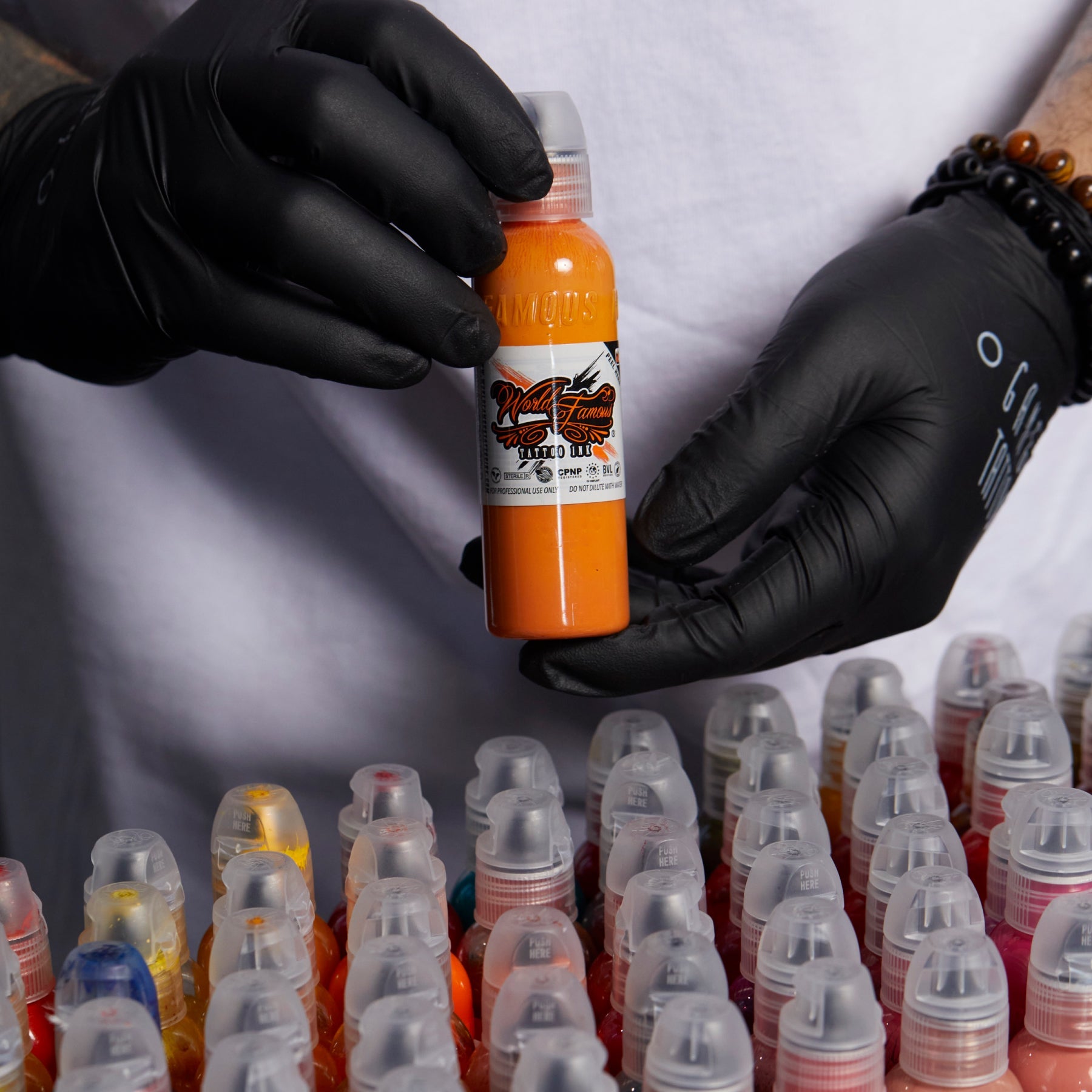 Gloved hands holding an orange World Famous Tattoo Ink bottle over a collection of ink bottles.