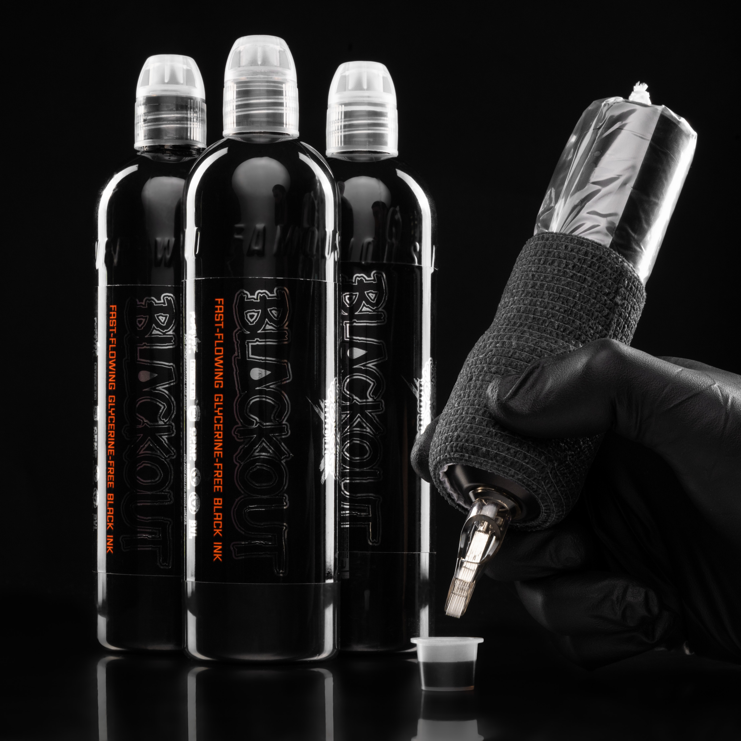 Three bottles of glycerin-free black tattoo ink standing behind a gloved hand holding a black-wrapped tattoo machine over an ink cap.
