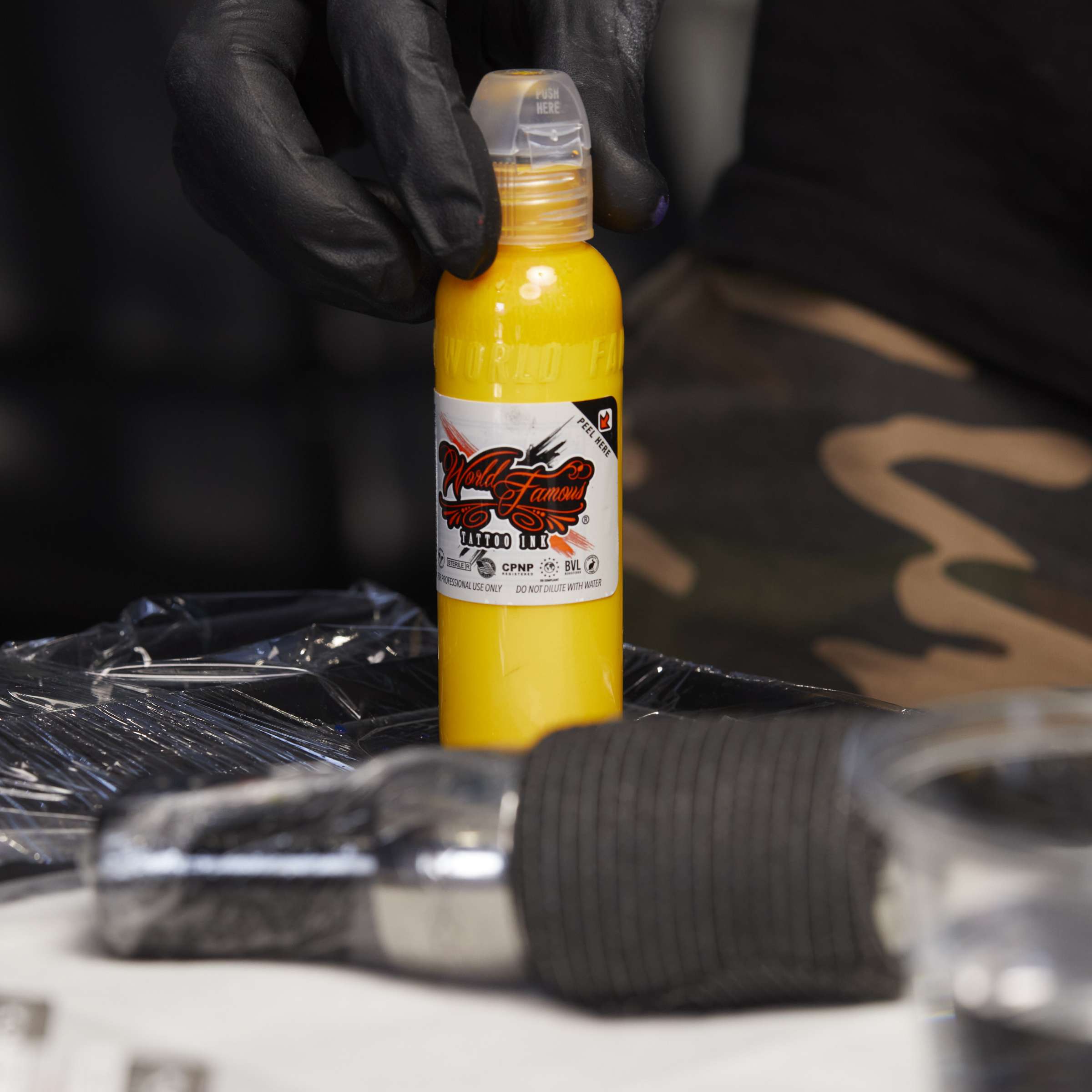A hand in a black glove picking up a bottle of bright yellow World Famous Tattoo Ink at an artist's workstation.