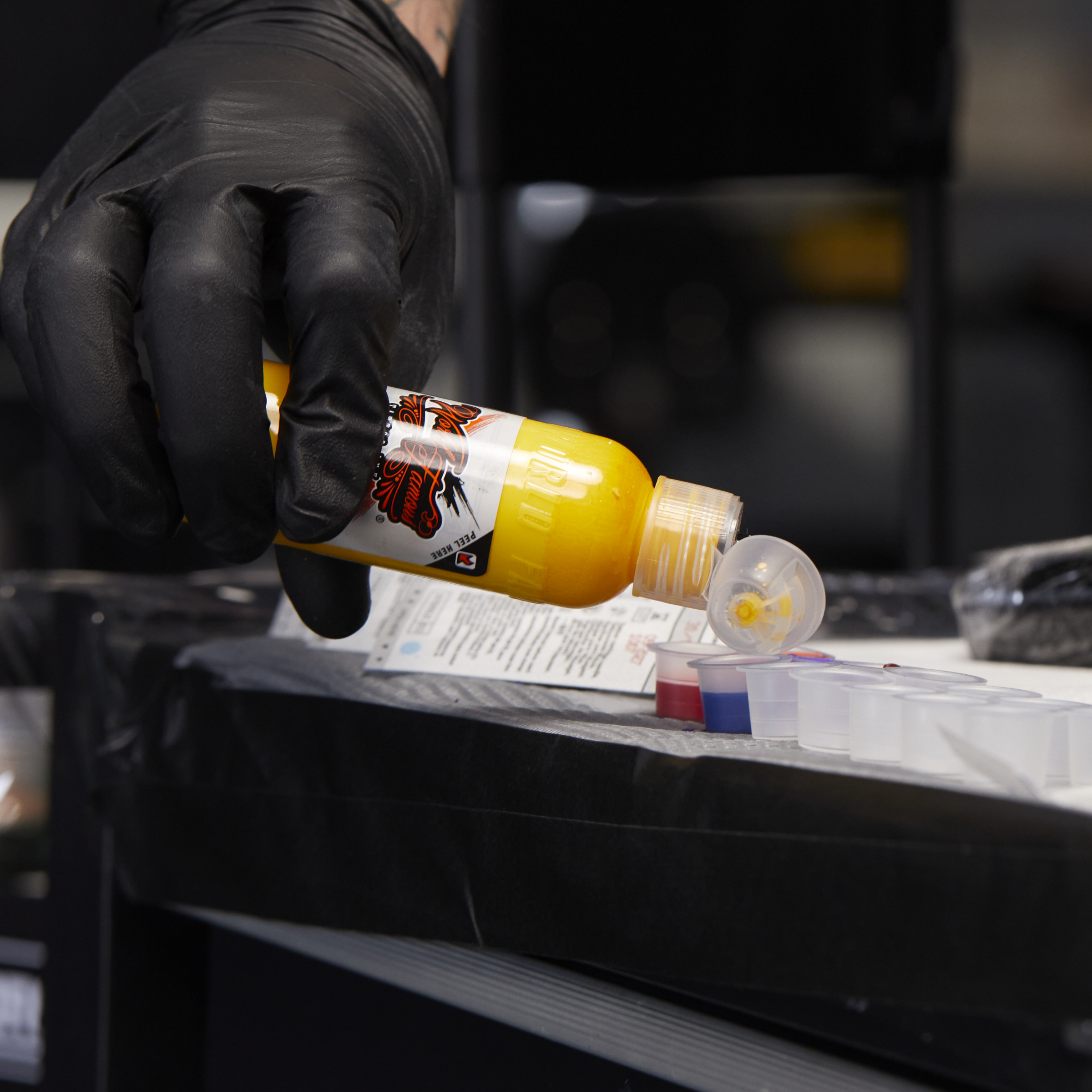 A tattoo artist wearing a black glove pours bright yellow World Famous Tattoo Ink into small plastic ink caps on a workstation.