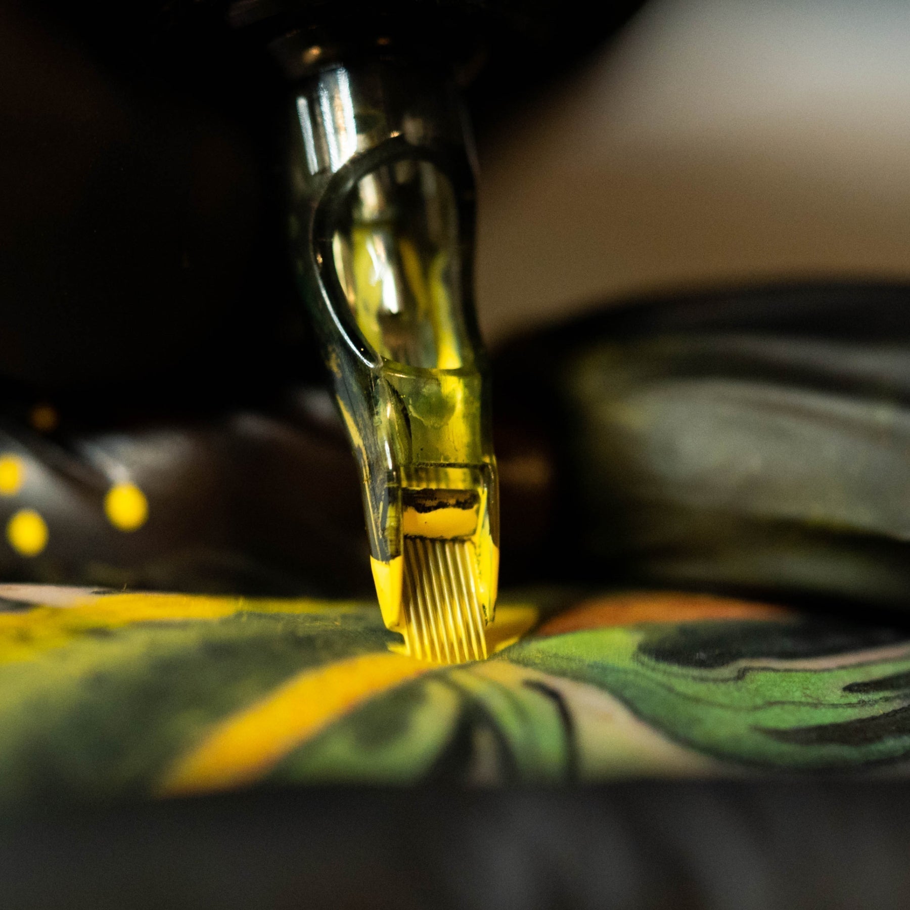 An extreme close-up of a tattooing needle cartridge loaded with bright yellow ink, precisely touching a skin surface covered in a colorful tattoo design.