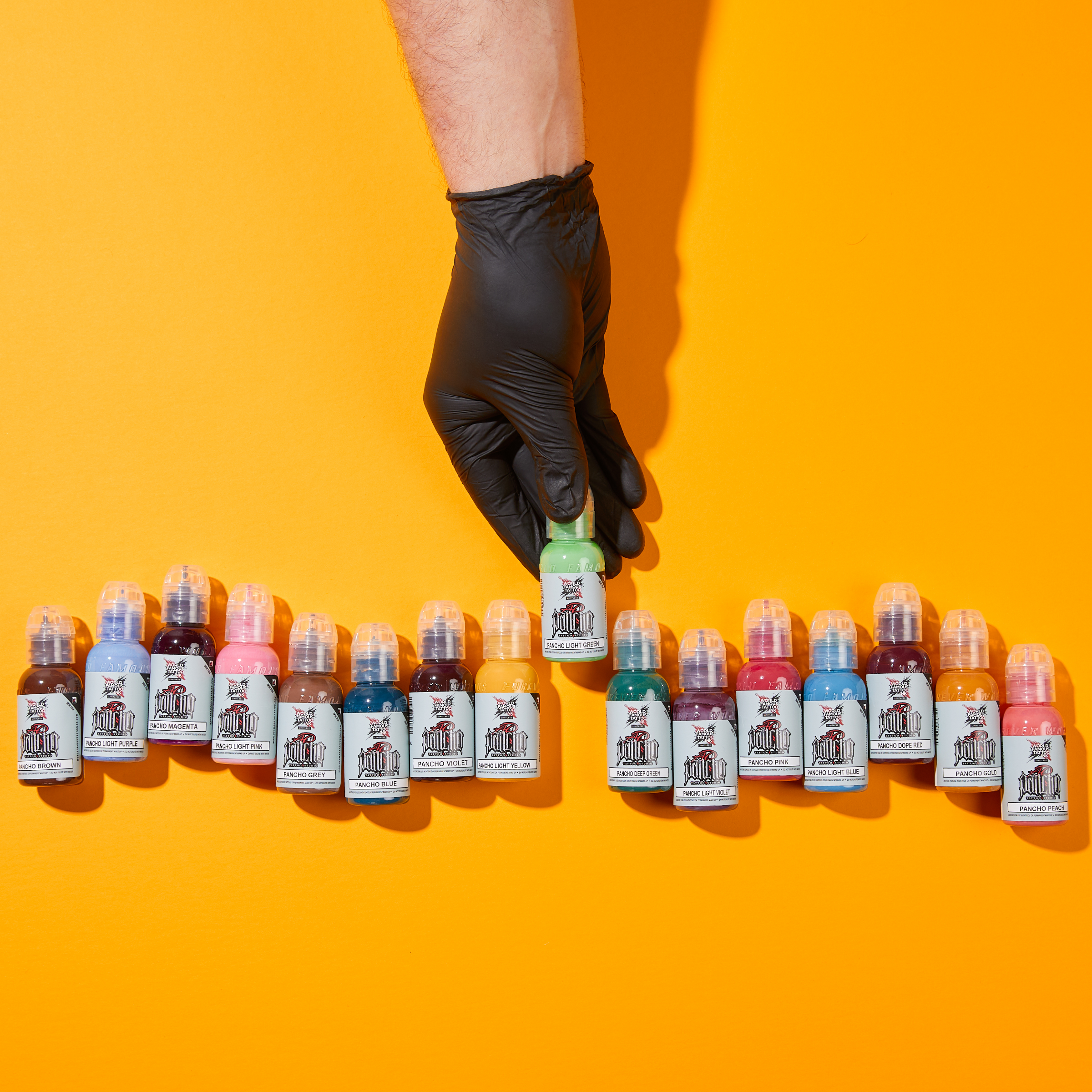 A gloved hand reaches for a bottle among a row of colourful supplies, highlighting professional tattoo ink formulation
