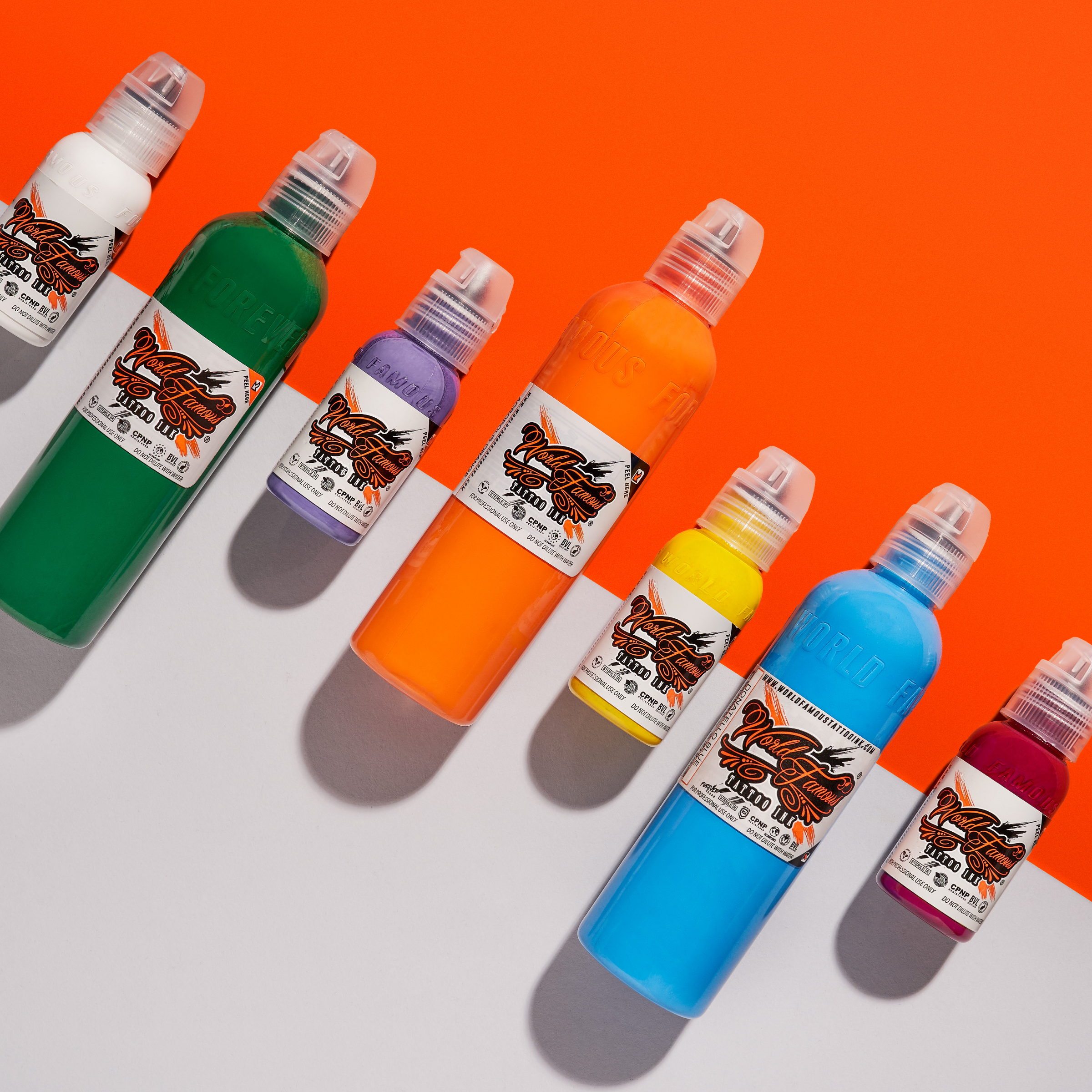 Seven multi-colored bottles of tattoo ink, including green, purple, orange, and blue, arranged in a diagonal line on a split orange and light gray background.