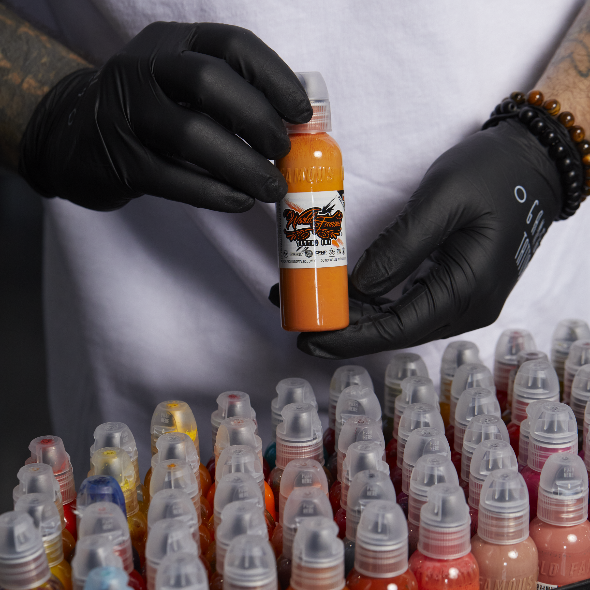 A tattoo artist wearing black gloves holds up a bottle of orange World Famous Tattoo Ink above a large collection of ink caps.